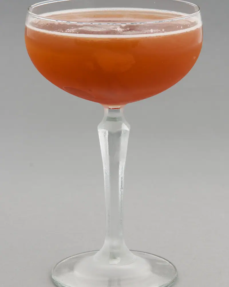 A Blood and Sand, reddish-orange, in a coupe glass against a plain grey background