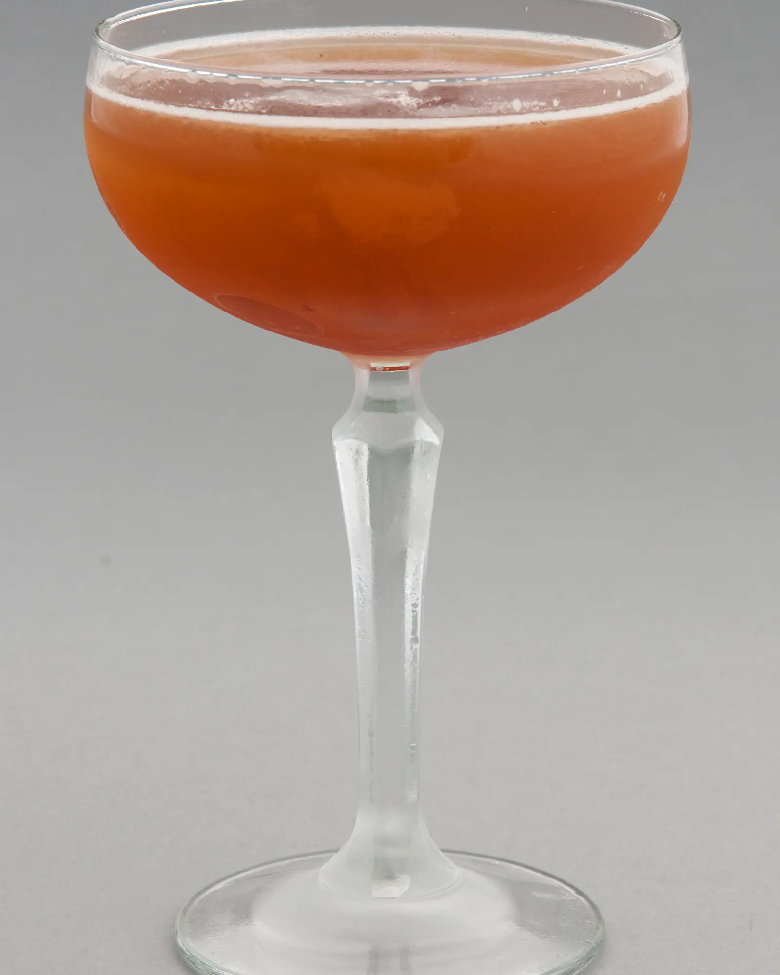 A Blood and Sand, reddish-orange, in a coupe glass against a plain grey background