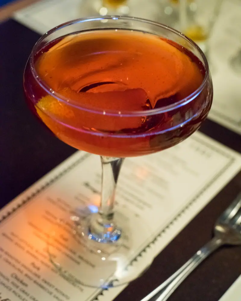A Boulevardier served up in a coupe, deep amber-red, with an orange twist resting against the rim.