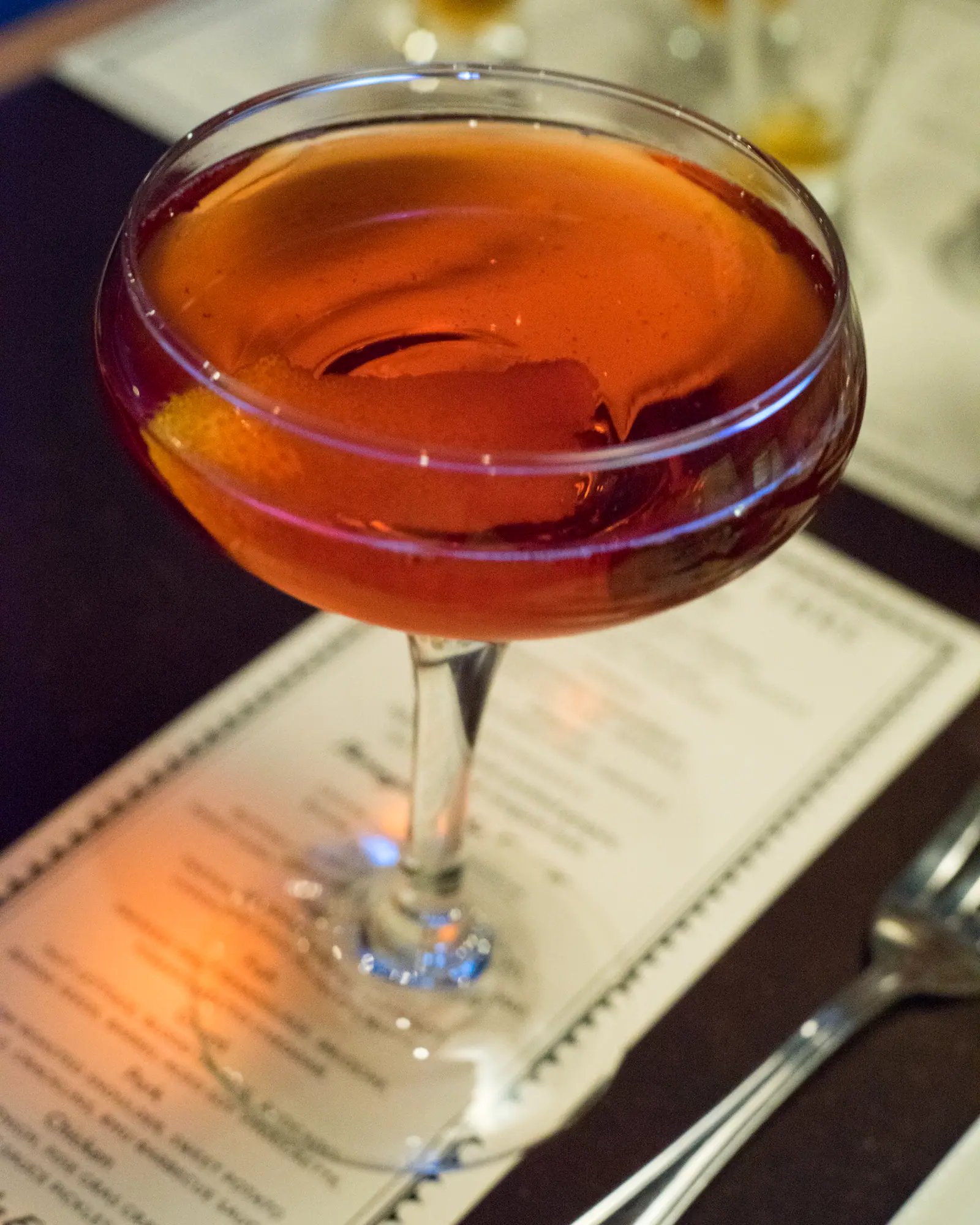 A Boulevardier served up in a coupe, deep amber-red, with an orange twist resting against the rim.