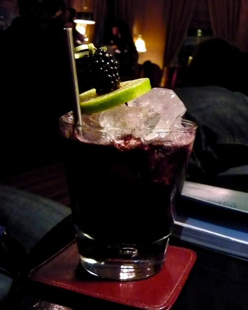 A Bramble cocktail in a rocks glass over crushed ice, deep purple-red, garnished with a blackberry and a lime wheel on a cocktail pick