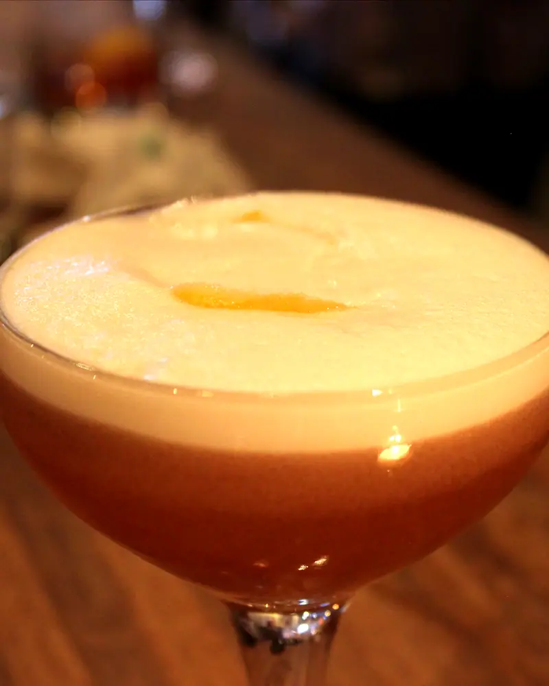 A Clover Club cocktail in a coupe, layered pink beneath a dense white foam, garnished with a raspberry on the surface