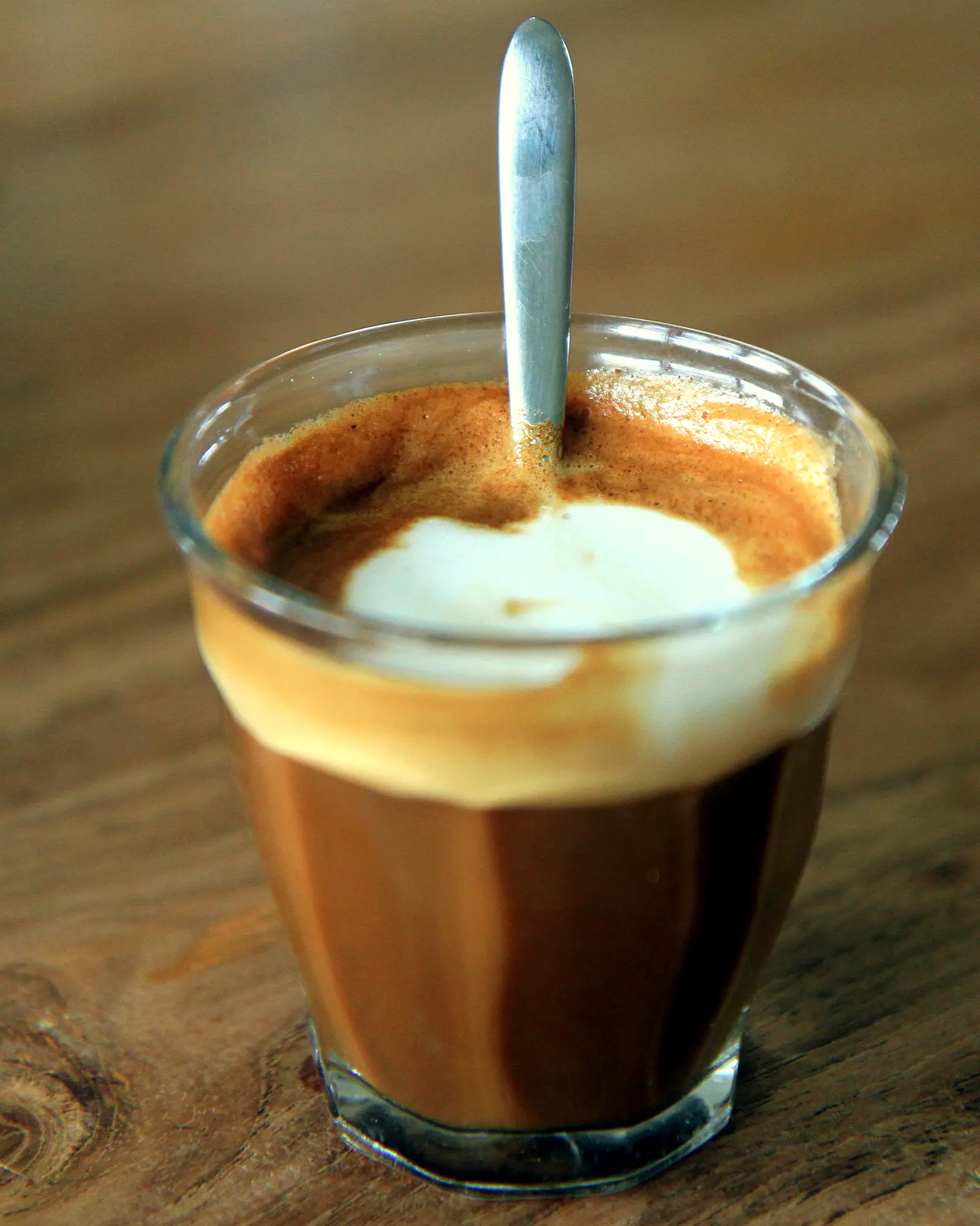 A cortado in a small glass, espresso cut with steamed milk, a spoon resting inside