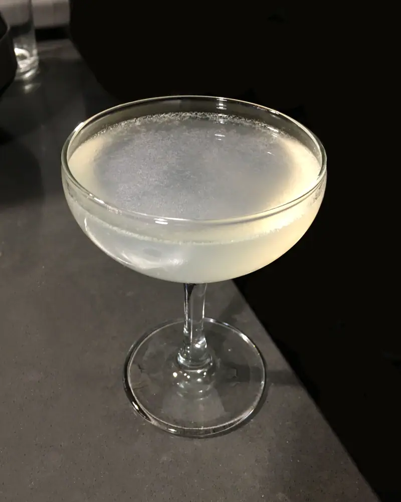 A Daiquiri served up in a coupe, pale and faintly cloudy under a thin white foam, on a dark grey counter