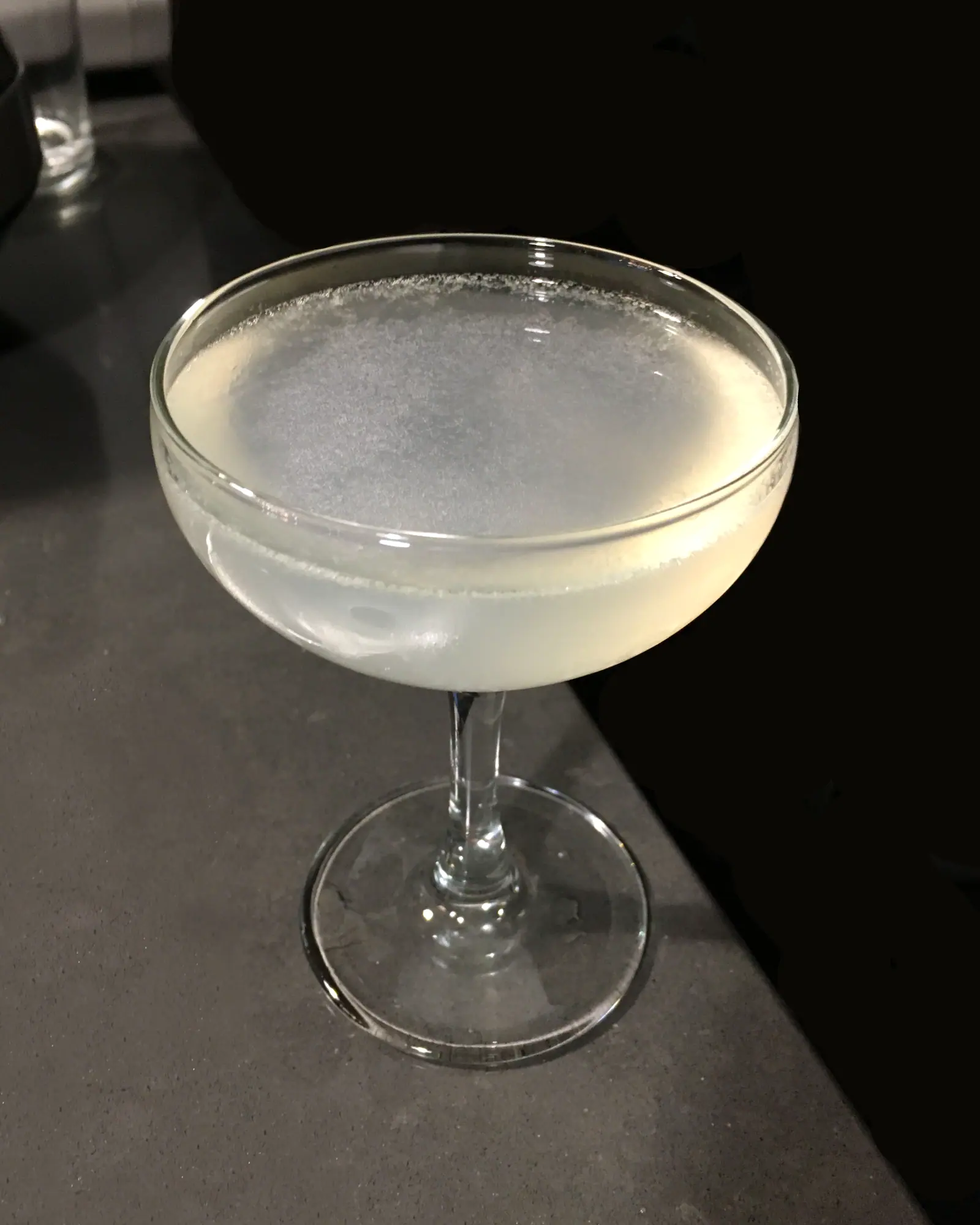 A Daiquiri served up in a coupe, pale and faintly cloudy under a thin white foam, on a dark grey counter