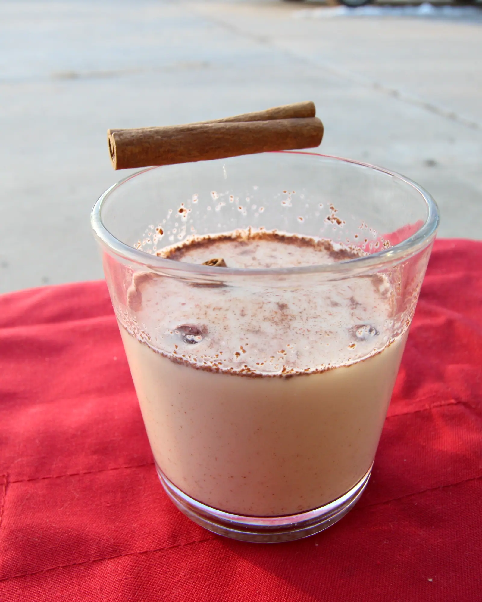 A glass of pale tan eggnog dusted with cinnamon, a cinnamon stick resting across the rim