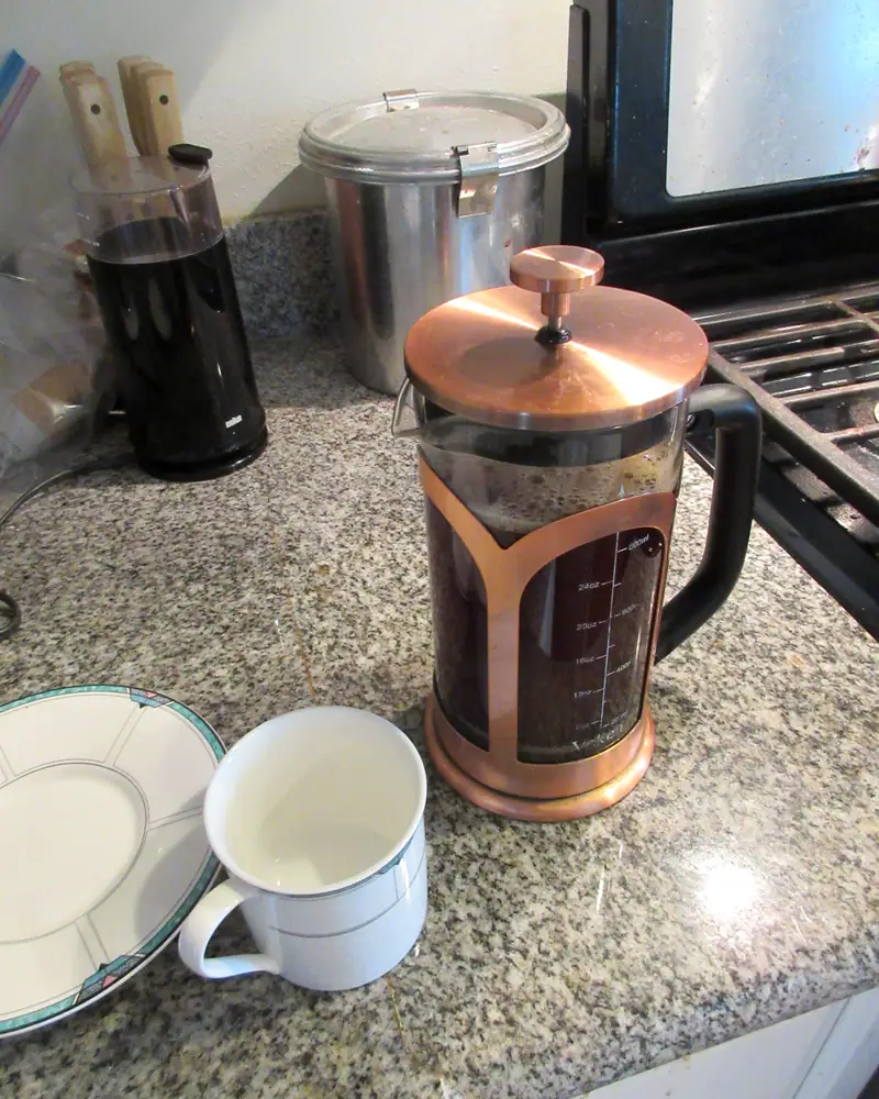 A copper-trimmed glass French press full of brewed coffee beside a white mug, on a kitchen counter