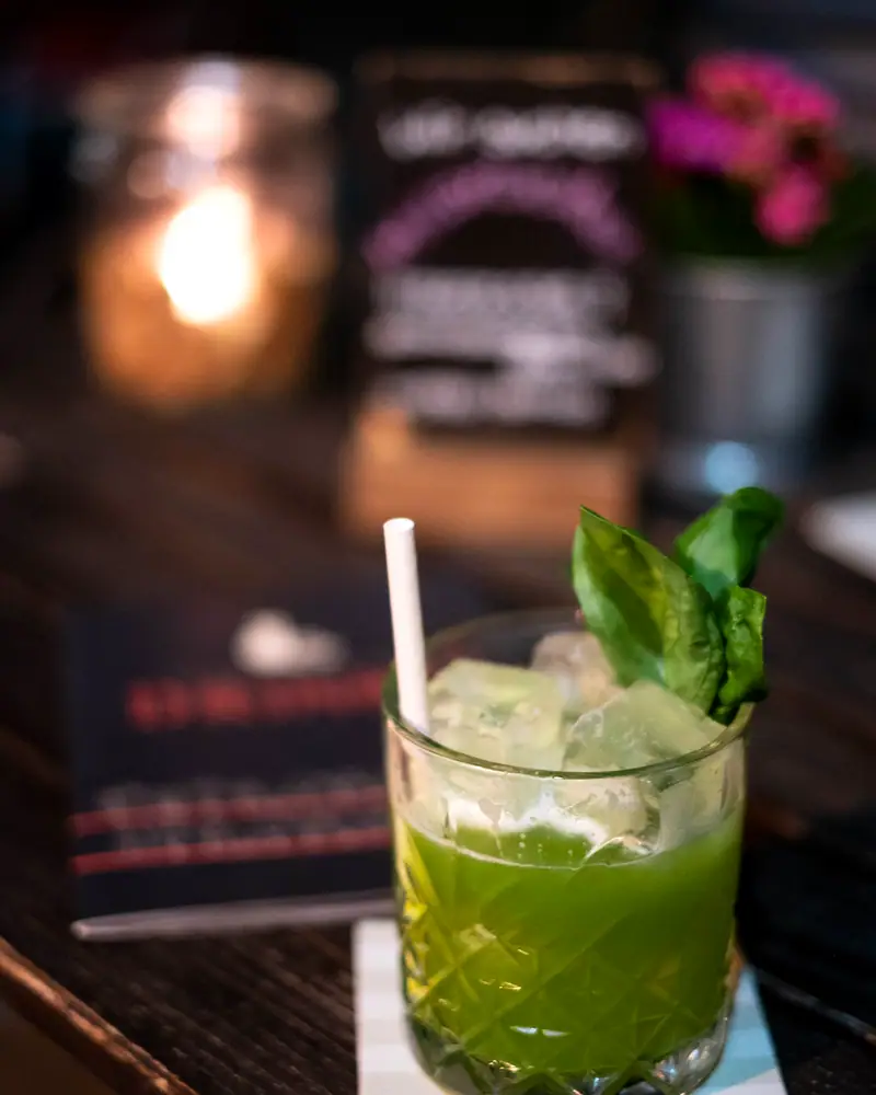 A Gin Basil Smash in a rocks glass over ice, pale green and topped with a fresh basil leaf, on a bar table with a candle behind it