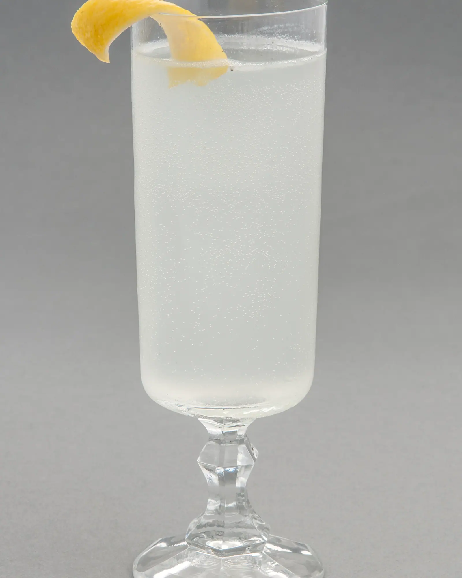 A clear, fizzy Gin Fizz in a tall stemmed glass, garnished with a curl of lemon peel