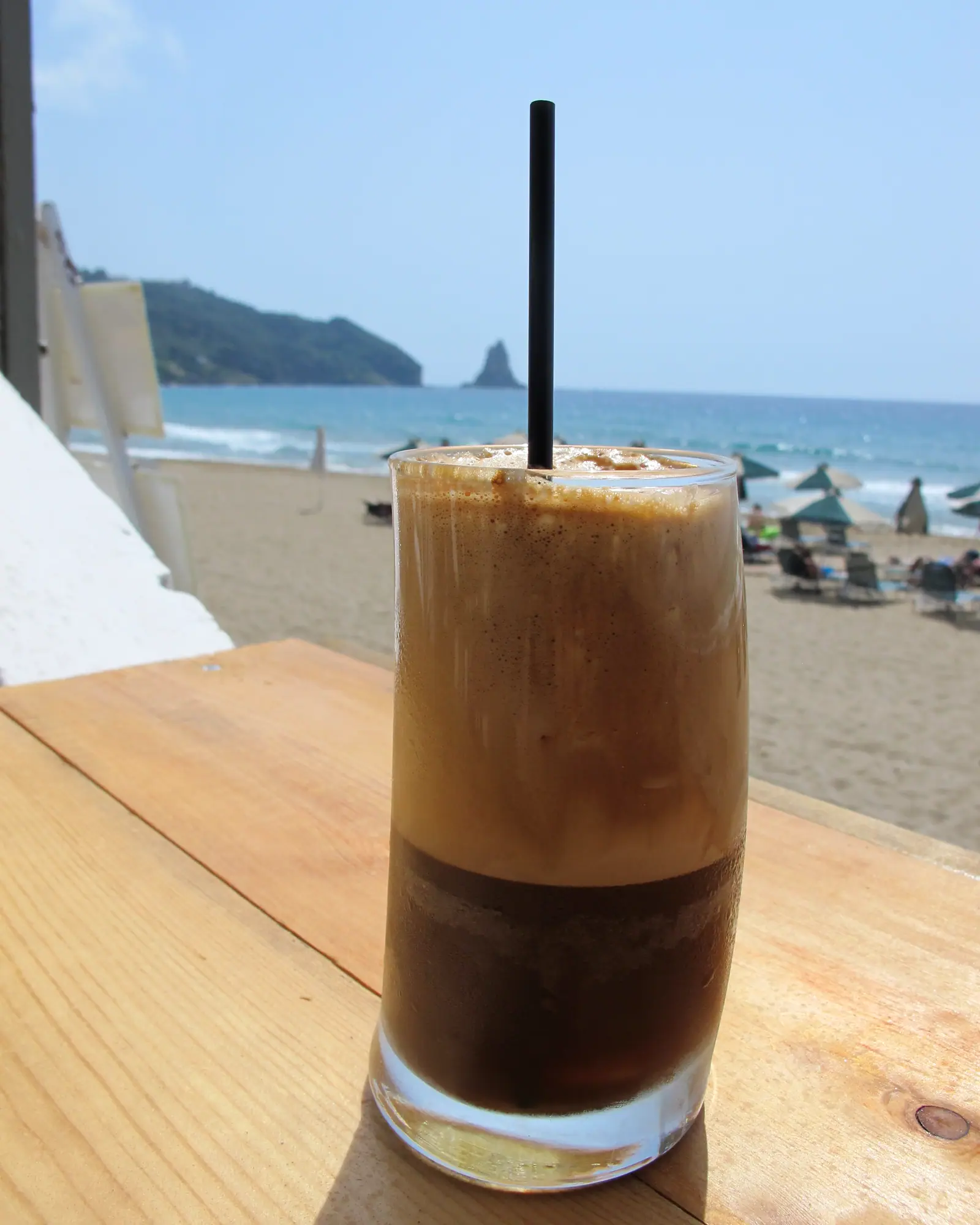 A tall glass of layered Greek frappé with dark foam over milky coffee, a straw, on a beachside table