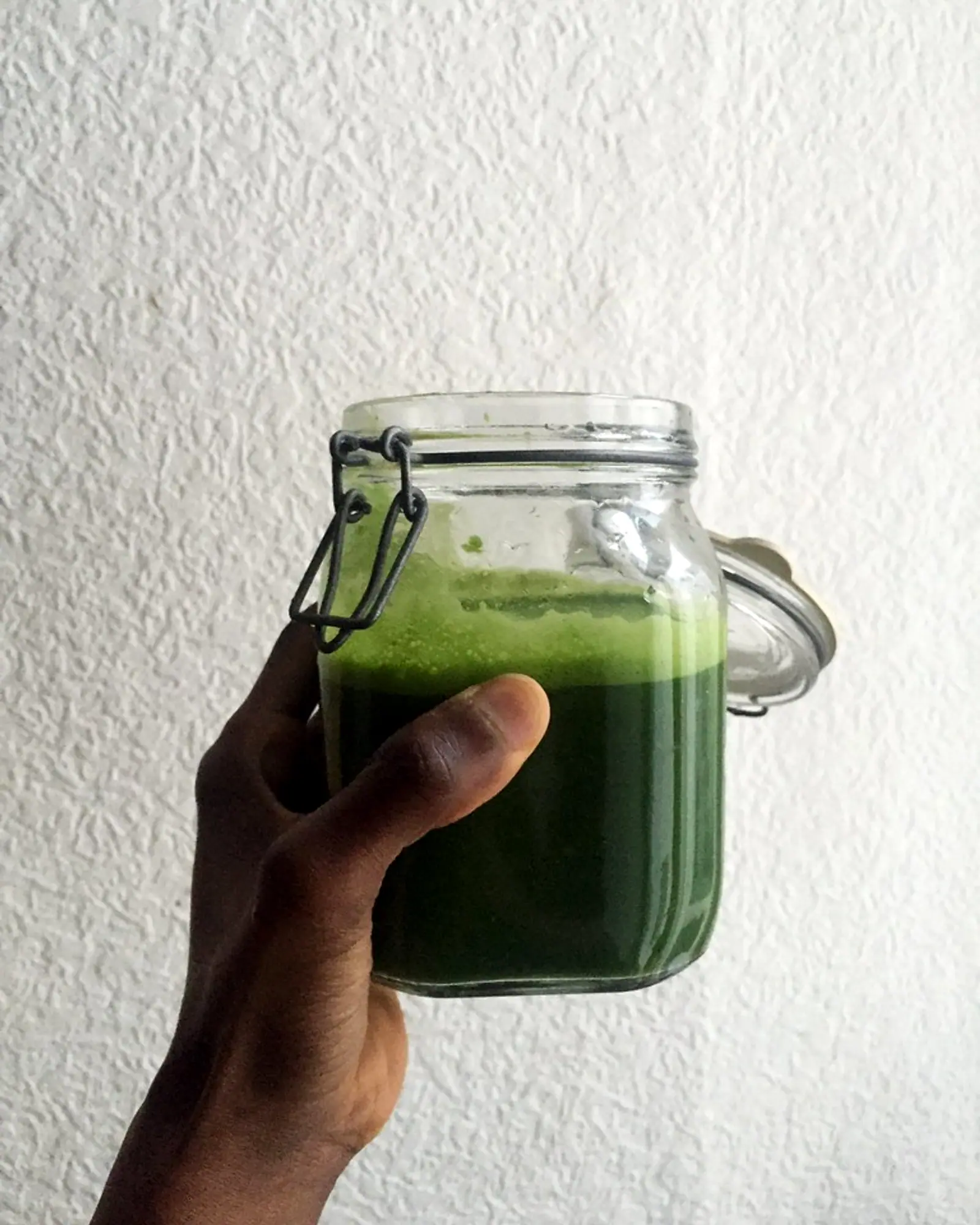 A hand holding up a glass jar of deep green cucumber-spinach juice against a white wall