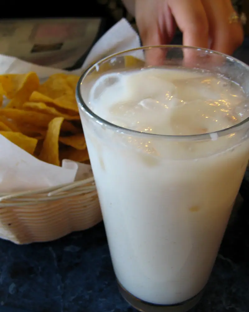 A tall glass of white, milky rice horchata over ice, beside a basket of tortilla chips