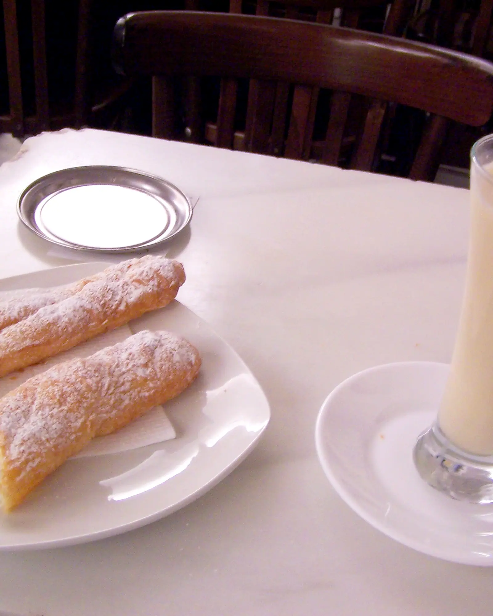 A tall glass of pale horchata de chufa beside a plate of sugar-dusted fartons on a café table