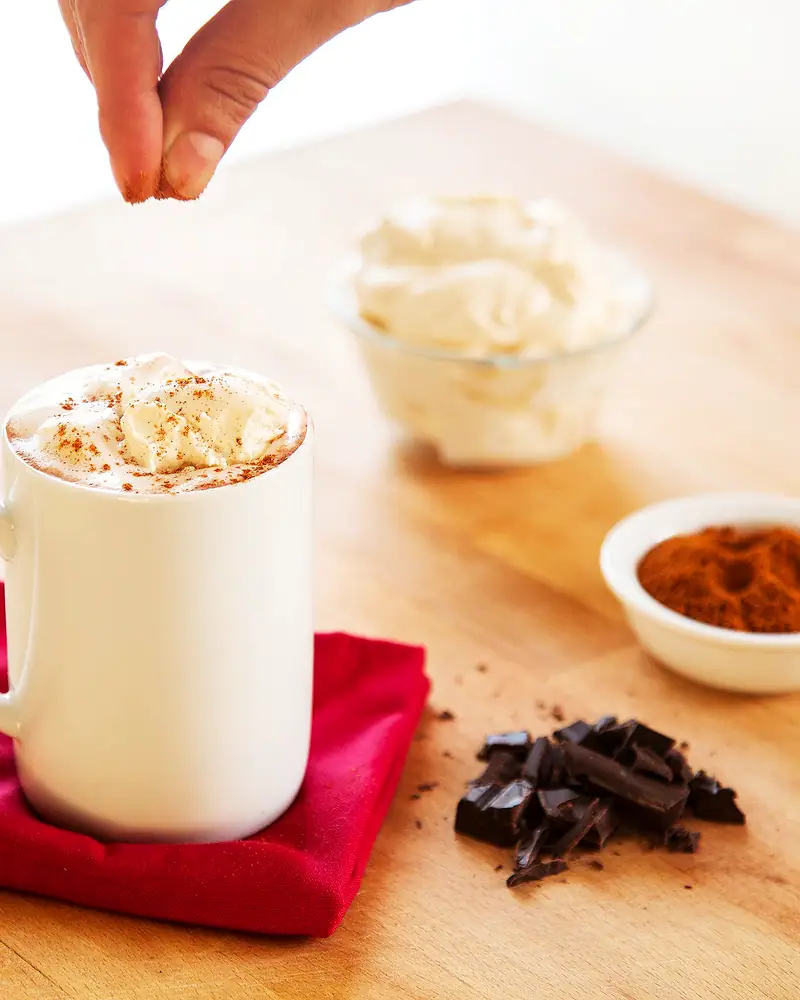 A white mug of hot chocolate topped with whipped cream, being dusted with spice, chocolate chunks and a bowl of cream beside it
