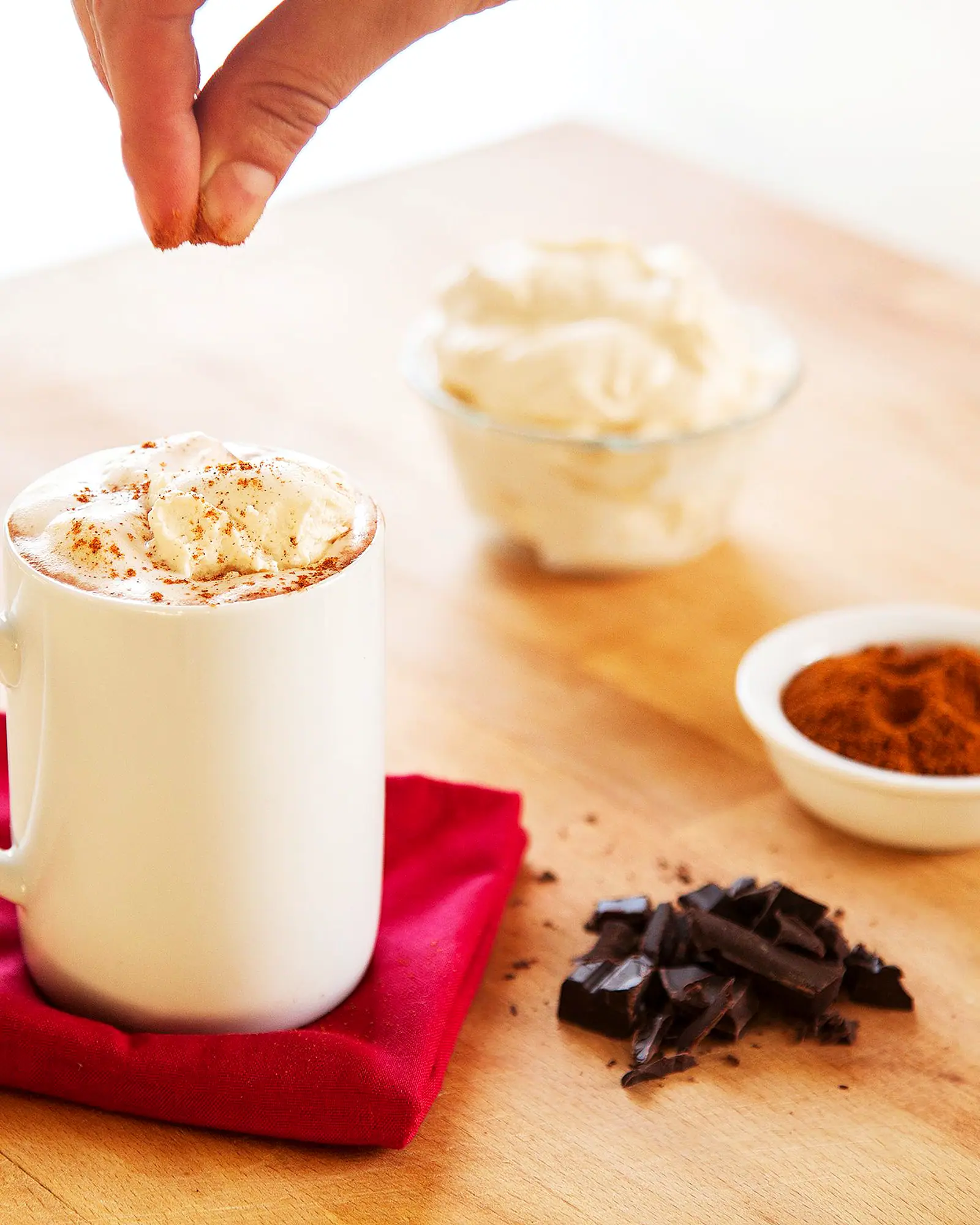 A white mug of hot chocolate topped with whipped cream, being dusted with spice, chocolate chunks and a bowl of cream beside it