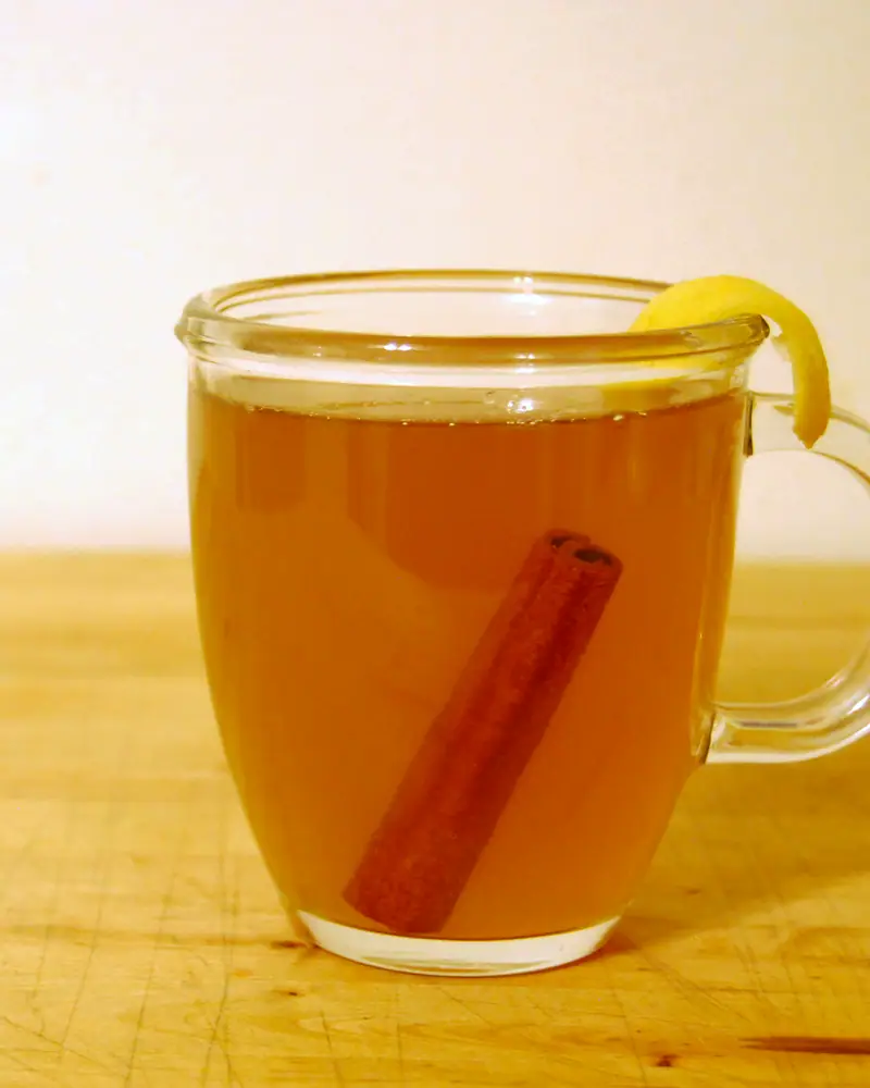 A glass mug of amber Hot Toddy with a cinnamon stick and a curl of lemon peel