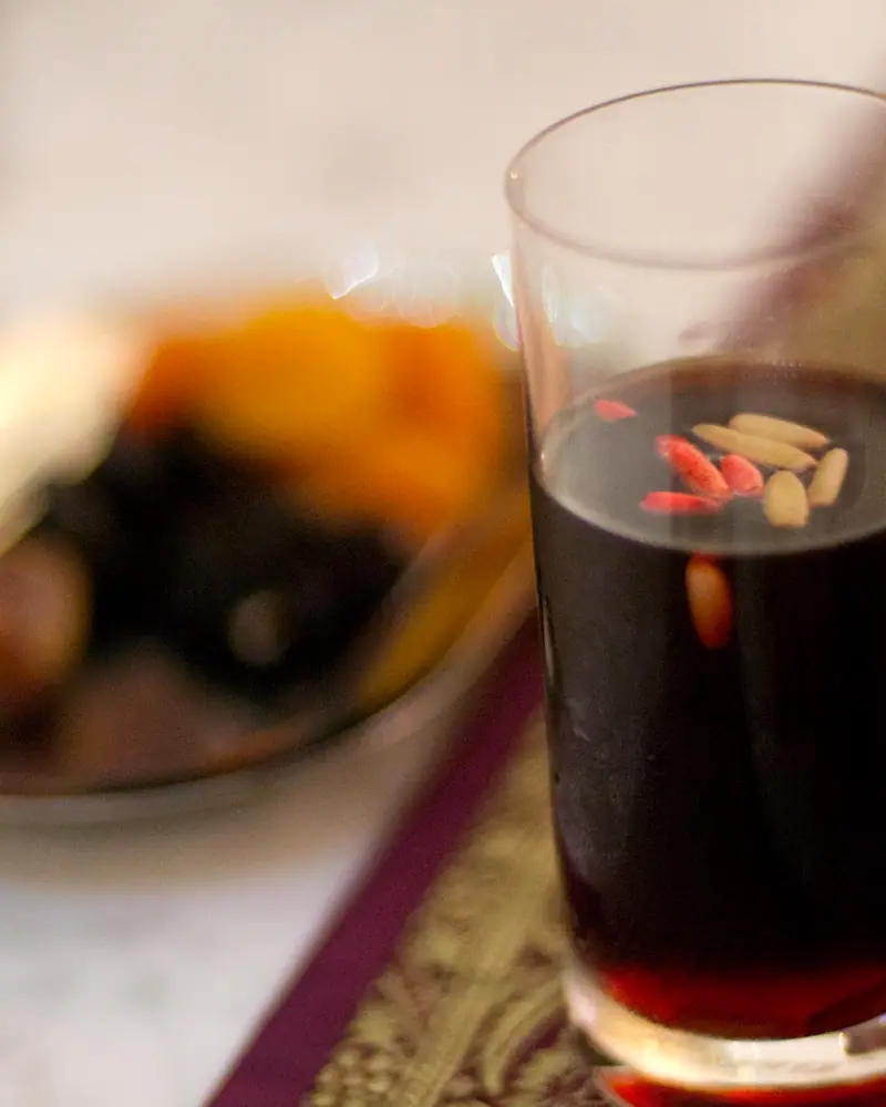 A glass of dark red jallab syrup drink with pine nuts floating on top