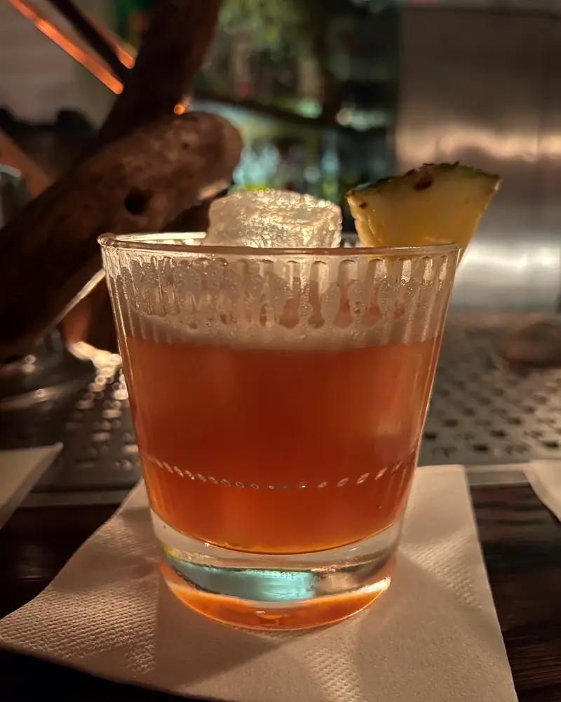 A Jungle Bird cocktail in a rocks glass over ice, amber-red in colour, garnished with a pineapple leaf