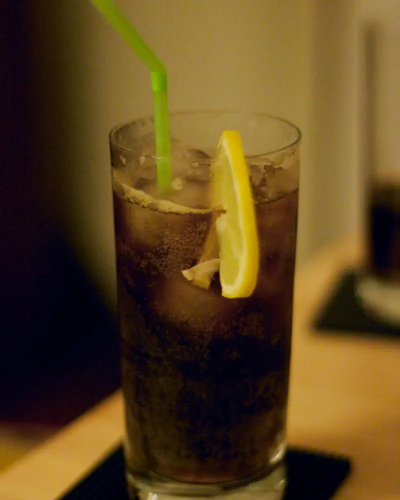 A Long Island Iced Tea in a tall glass over ice, garnished with a lemon wheel and a green straw
