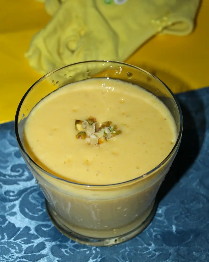 A glass of pale orange mango lassi topped with chopped pistachio