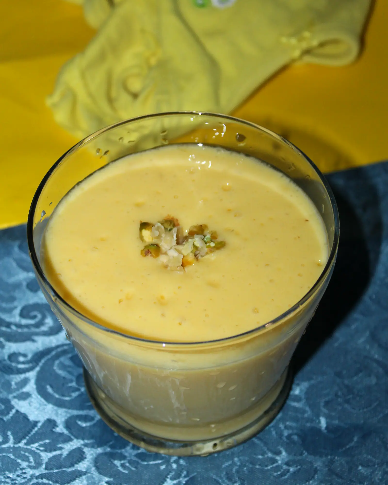 A glass of pale orange mango lassi topped with chopped pistachio