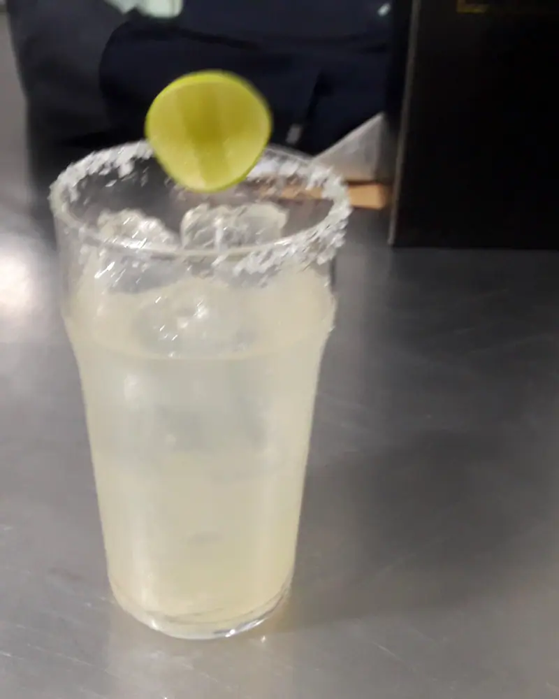 A pale, salt-rimmed Margarita over ice in a tall glass, garnished with a lime wheel