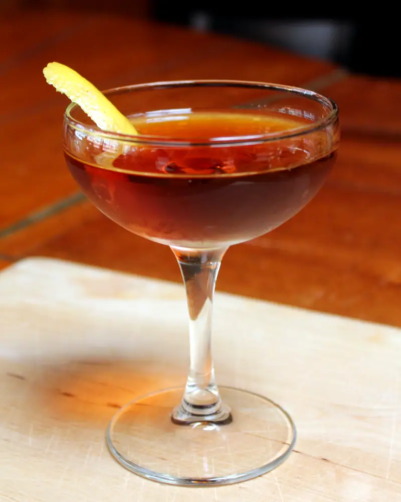 A Martinez served up in a coupe, deep amber-red, with a citrus twist resting on the rim.