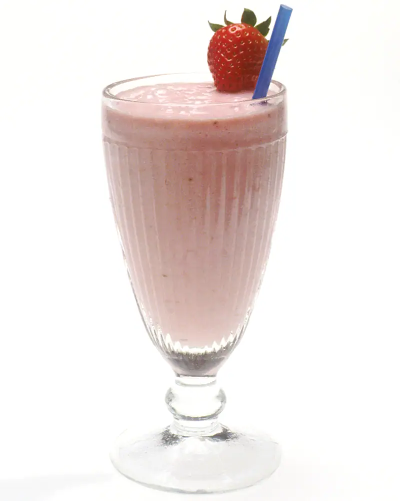 A pink strawberry milkshake in a fluted glass with a straw and a fresh strawberry garnish