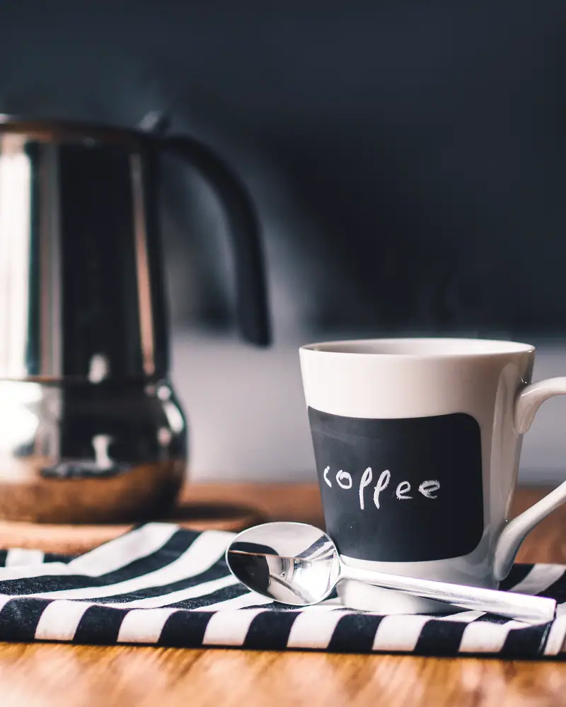 A cup of dark coffee beside a stovetop moka pot, on a striped cloth