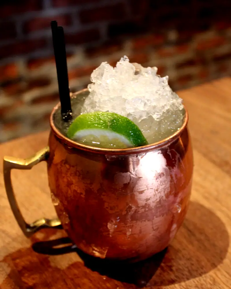 A Moscow Mule in a copper mug packed with crushed ice, garnished with a lime wheel and a black straw, on a wooden bar counter