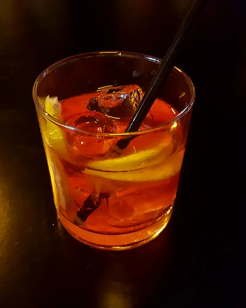 A Negroni in a rocks glass over ice, with a slice of orange and a stirrer, lit warmly against a dark bar