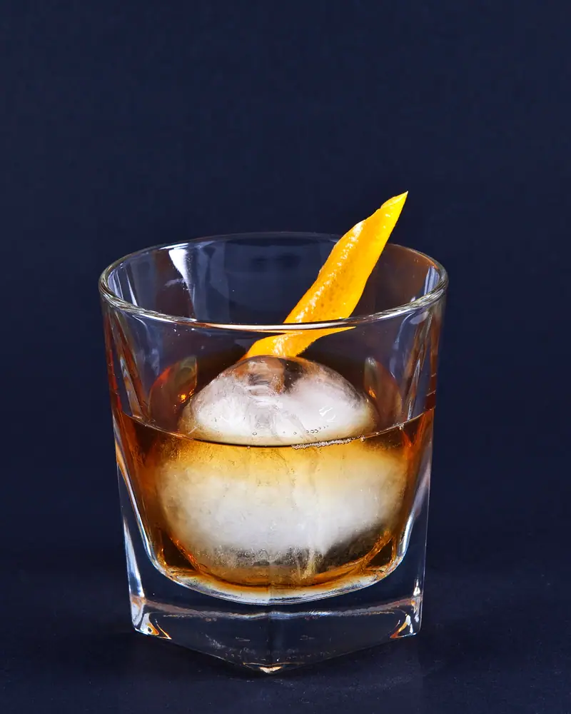 An Old Fashioned in a rocks glass, amber and clear, with a single large round ice cube and an orange twist standing against the rim