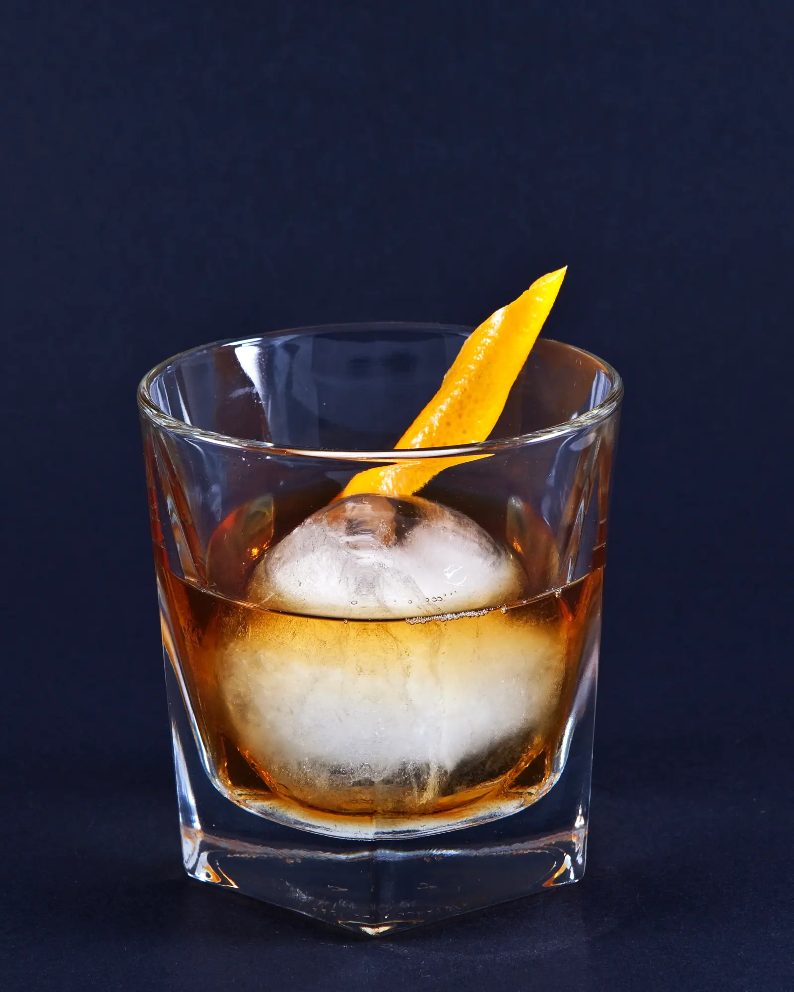 An Old Fashioned in a rocks glass, amber and clear, with a single large round ice cube and an orange twist standing against the rim