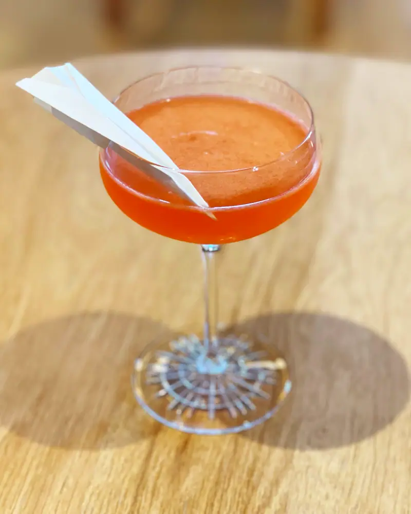 A Paper Plane cocktail served up in a coupe, orange-red in colour, garnished with a folded paper airplane