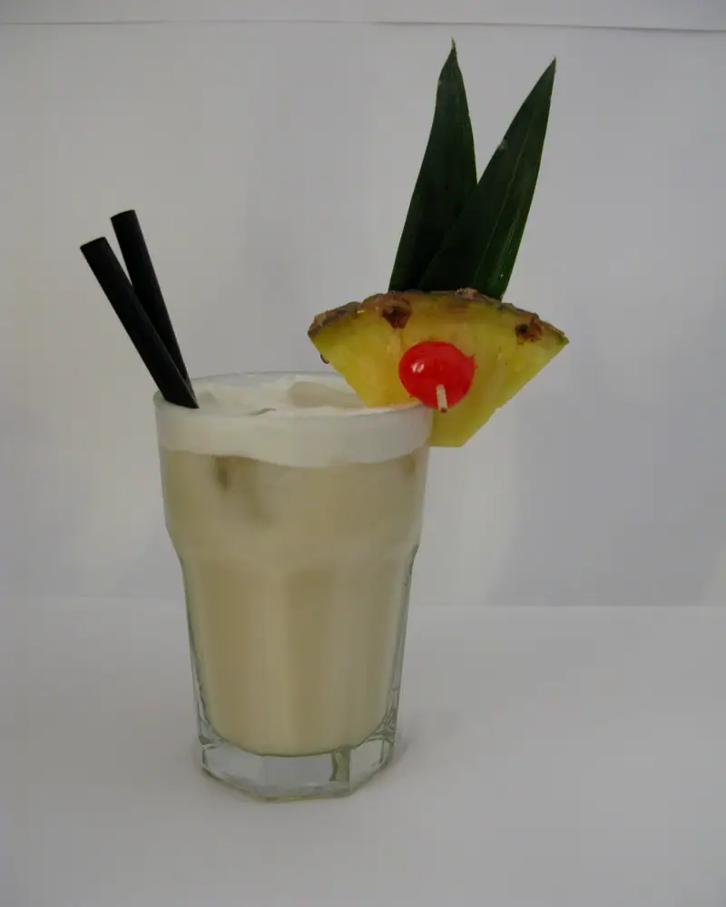 A Pina Colada served in a glass with a pineapple wedge, cherry and straws