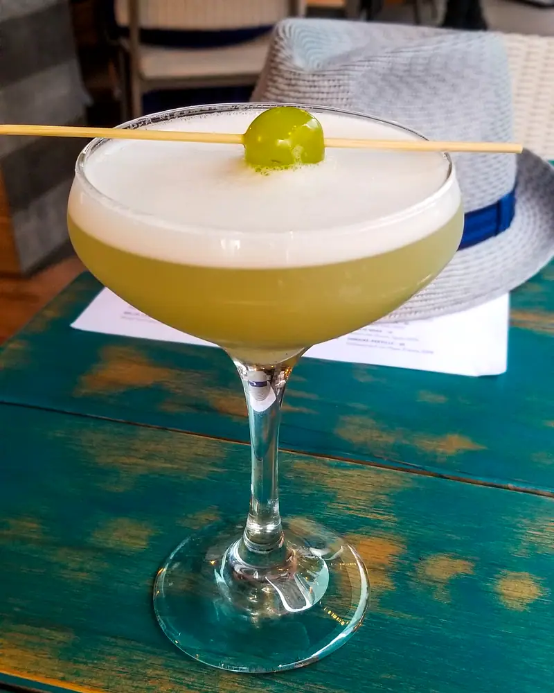A frothy pale yellow Pisco Sour served up in a coupe, garnished with a grape on a pick