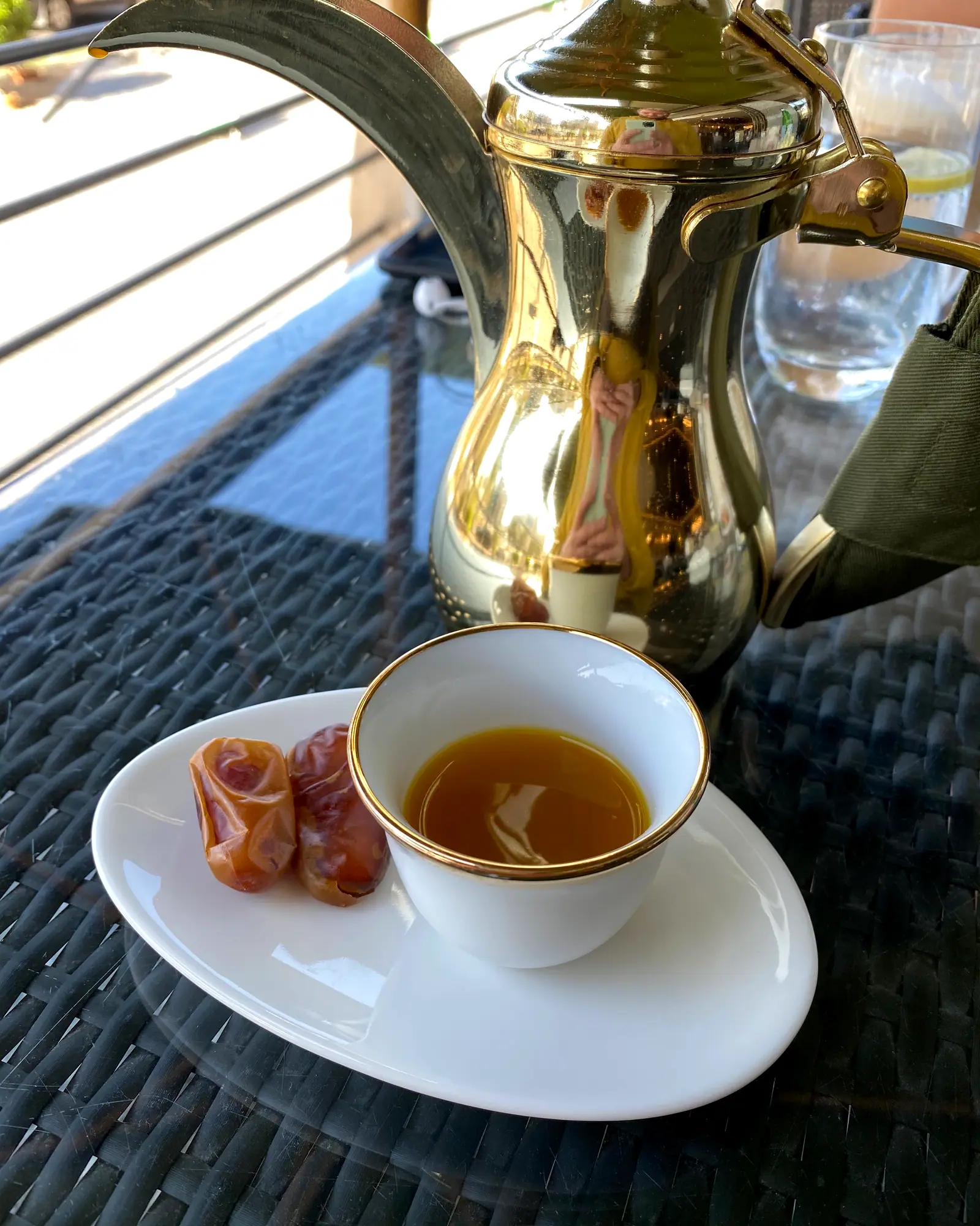 A small white cup of golden-brown qahwa on a saucer with dates, beside a brass dallah coffee pot