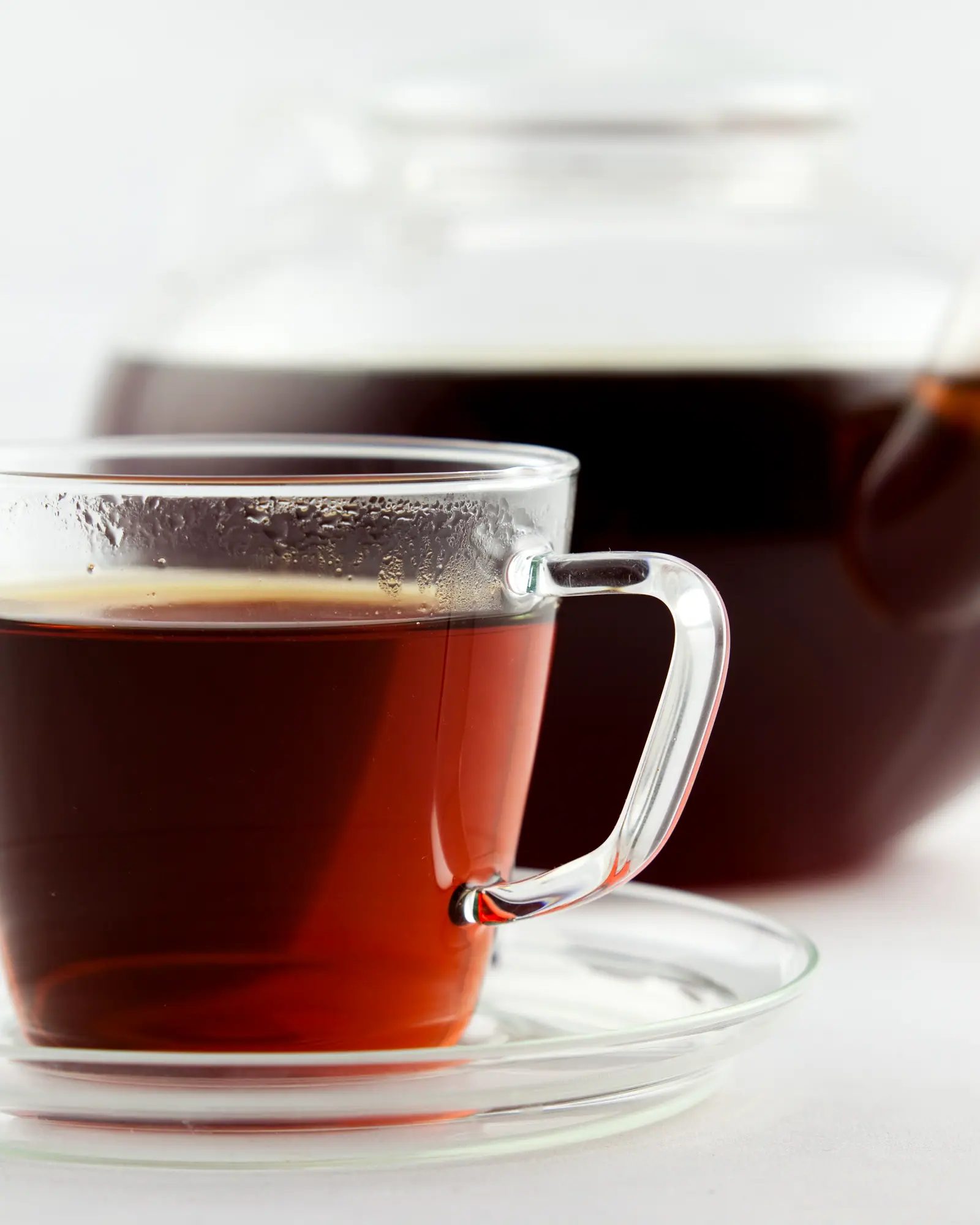A glass cup of freshly brewed rooibos tea, deep amber-red, with a glass teapot behind it