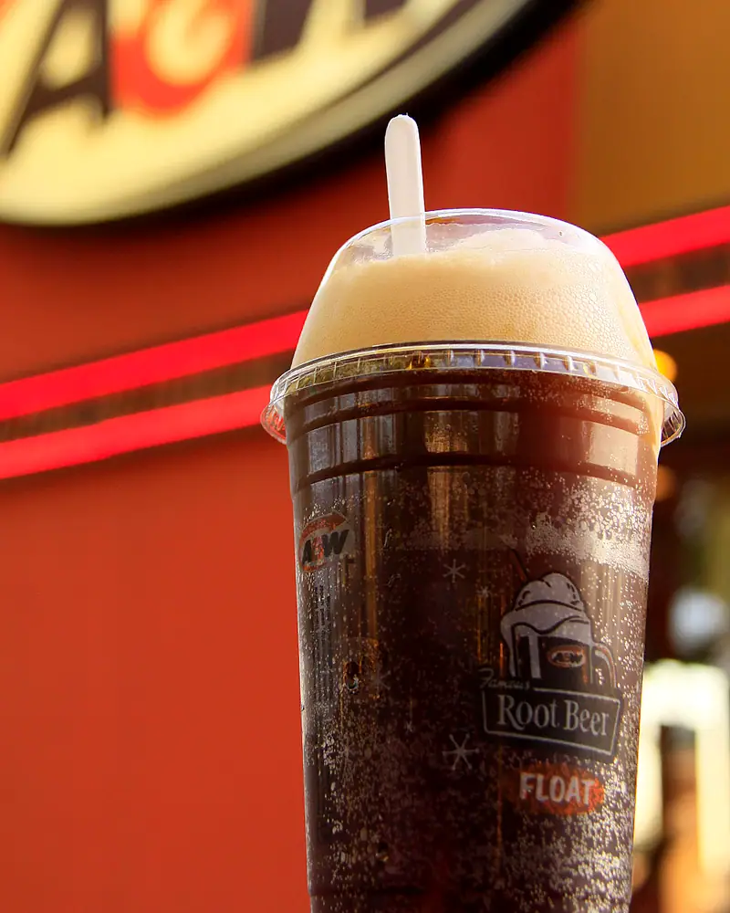A root beer float in a clear cup printed 'Root Beer Float', dark root beer topped with white foam and a straw, an A&W sign visible behind