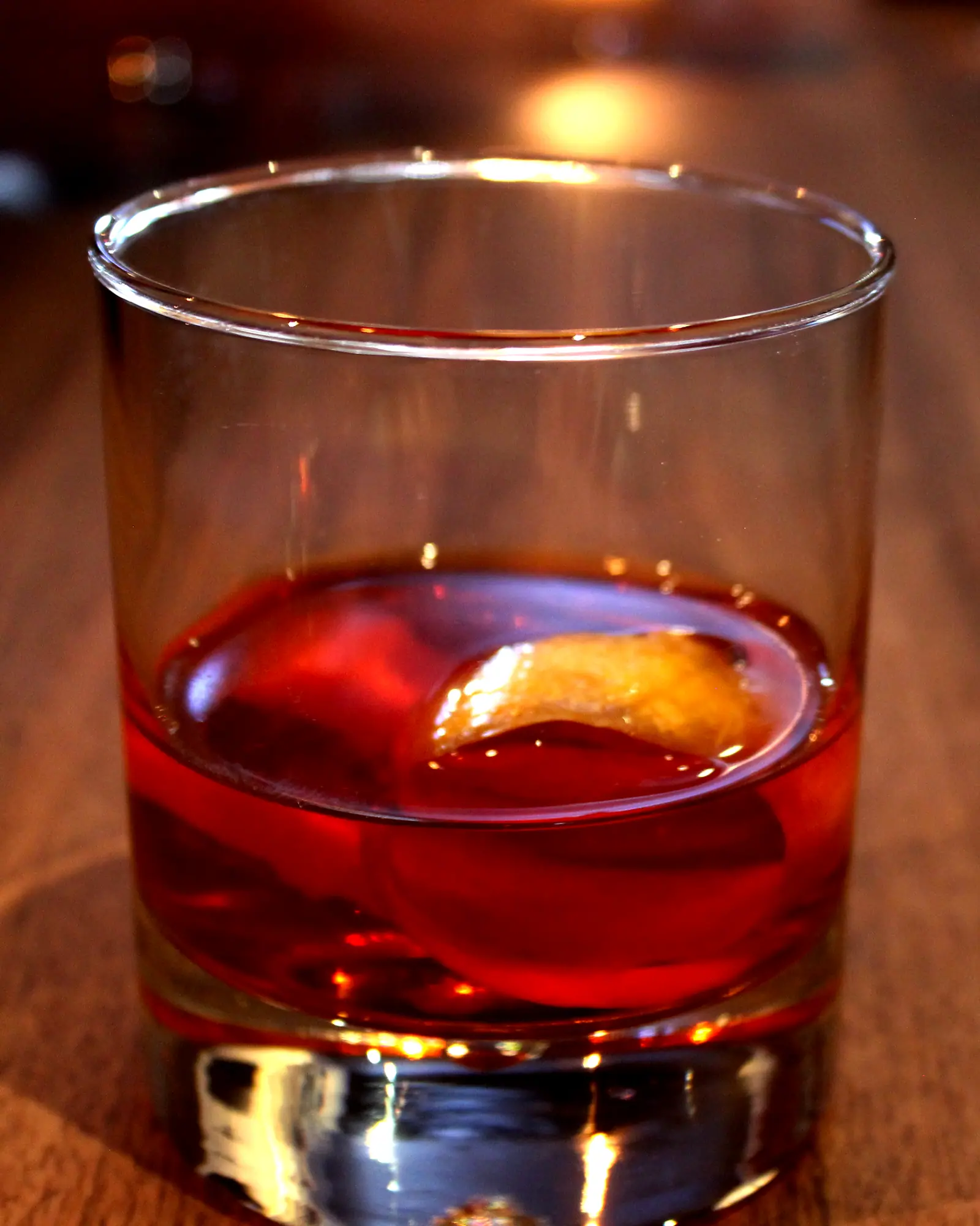 A Sazerac served up in a rocks glass with no ice, deep amber-red, with a lemon peel resting on the surface