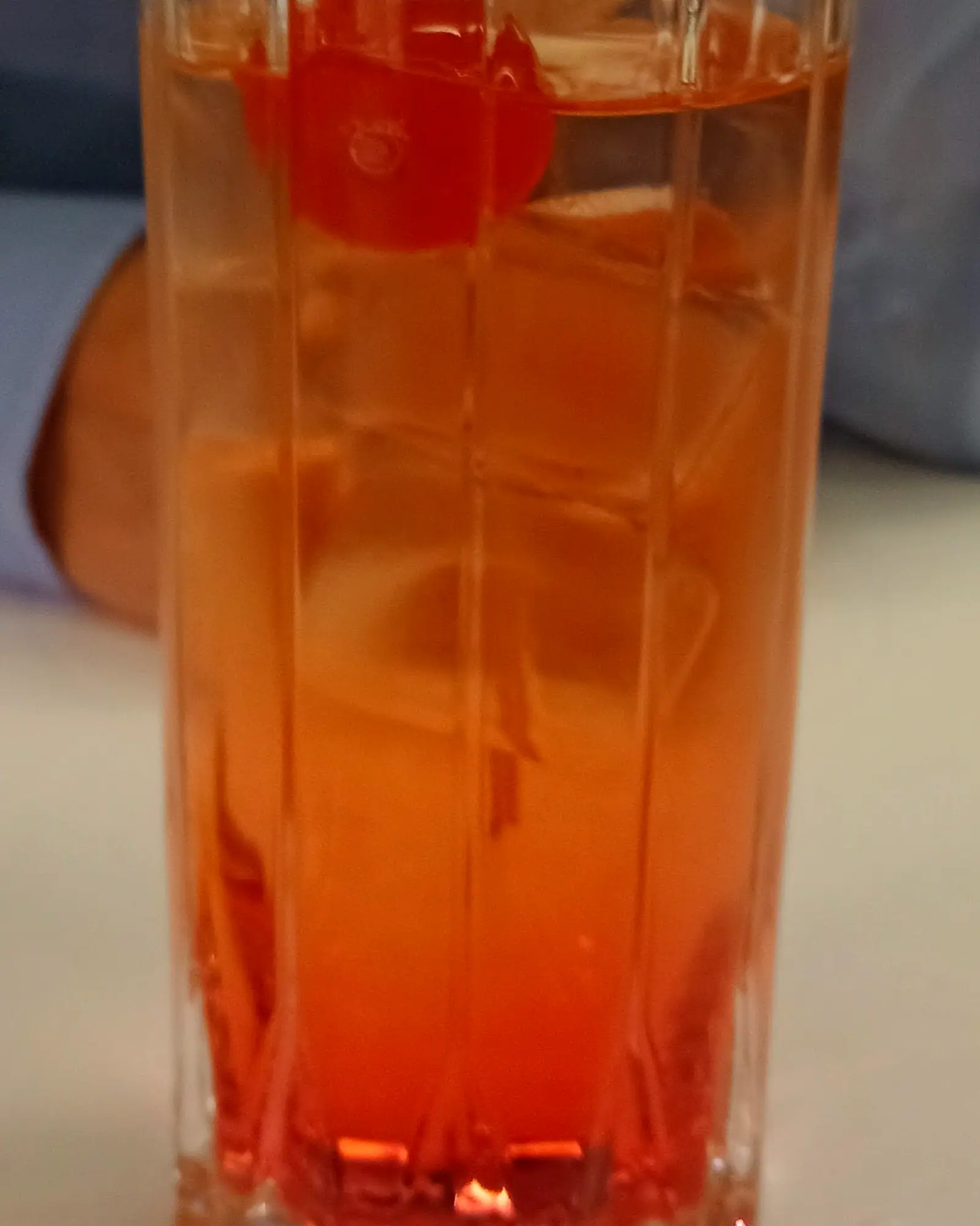 A Shirley Temple in a tall glass over ice, orange ginger ale with red grenadine settled at the bottom, garnished with a maraschino cherry