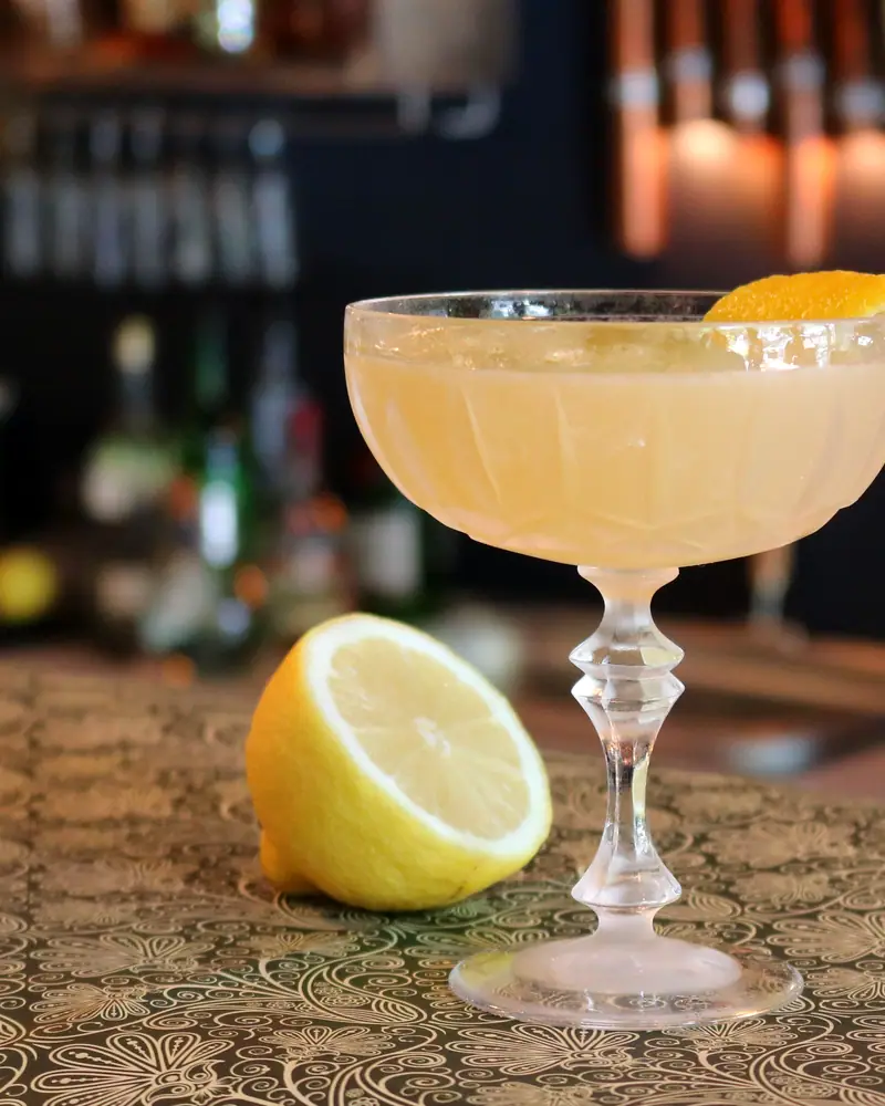A sugar-rimmed Sidecar served up in a coupe, amber-gold, beside a lemon half