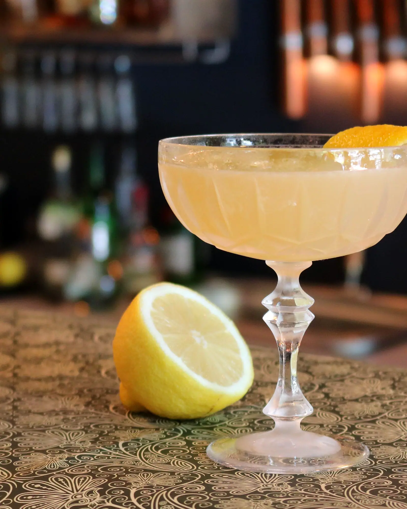 A sugar-rimmed Sidecar served up in a coupe, amber-gold, beside a lemon half
