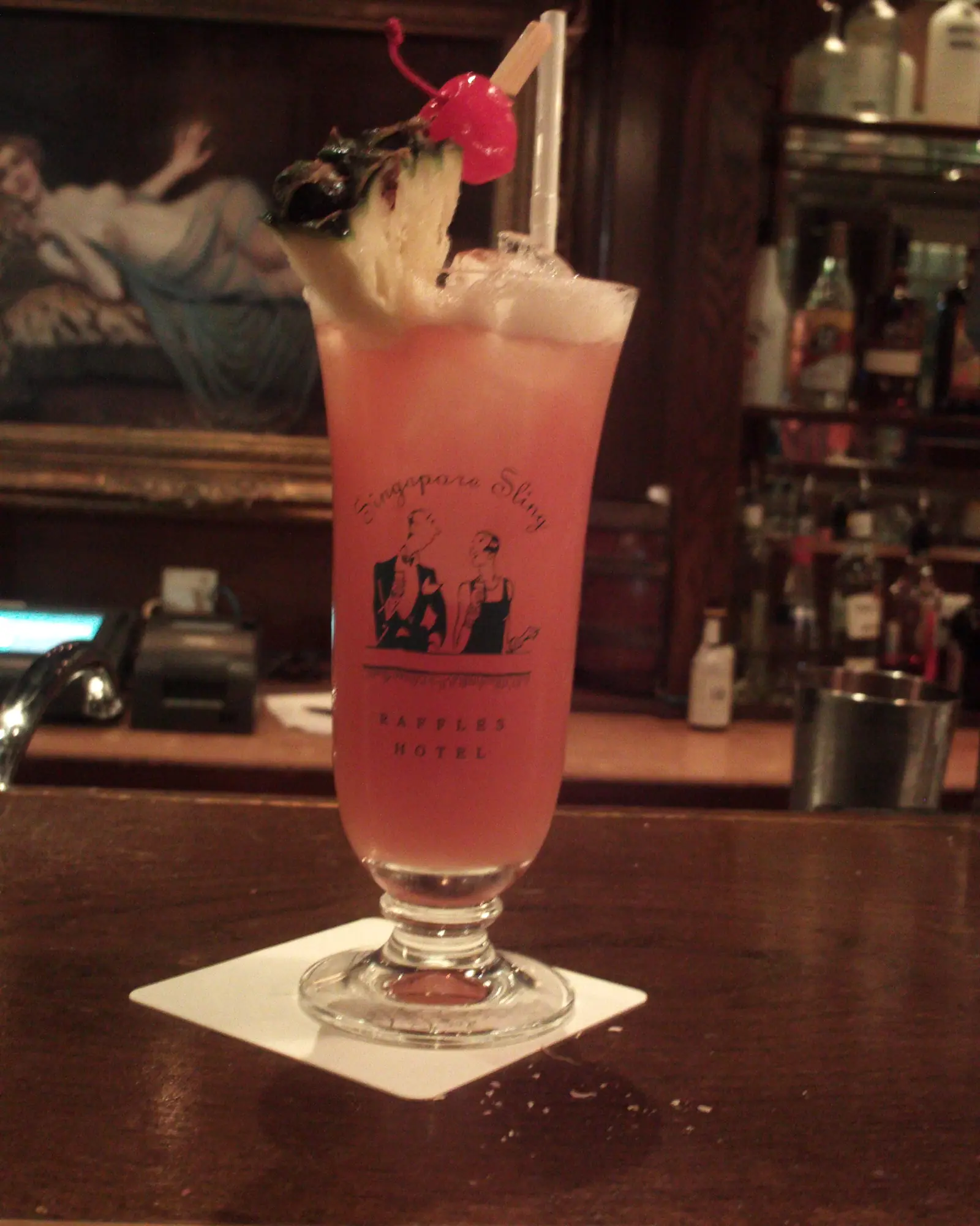 A Singapore Sling in a footed glass on a wooden bar top, its pink-red drink topped with foam and garnished with a pineapple wedge, mint sprig and a cherry on a pick, dark bar shelving blurred behind it