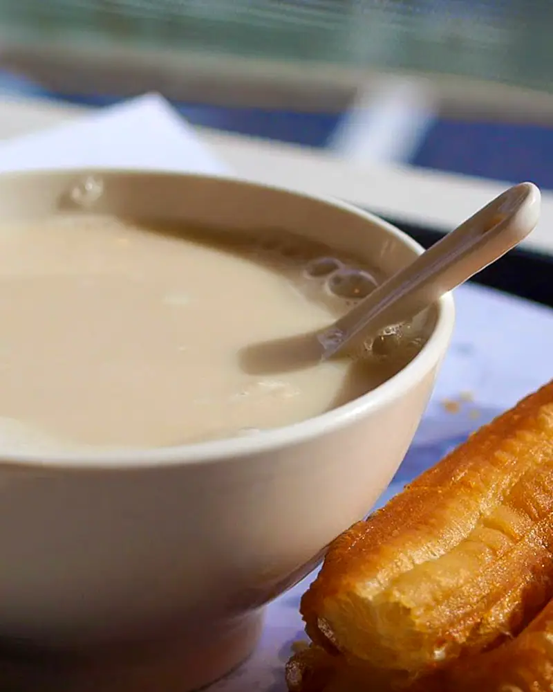 A bowl of warm plain soy milk (doujiang) with a spoon, served alongside a fried dough stick