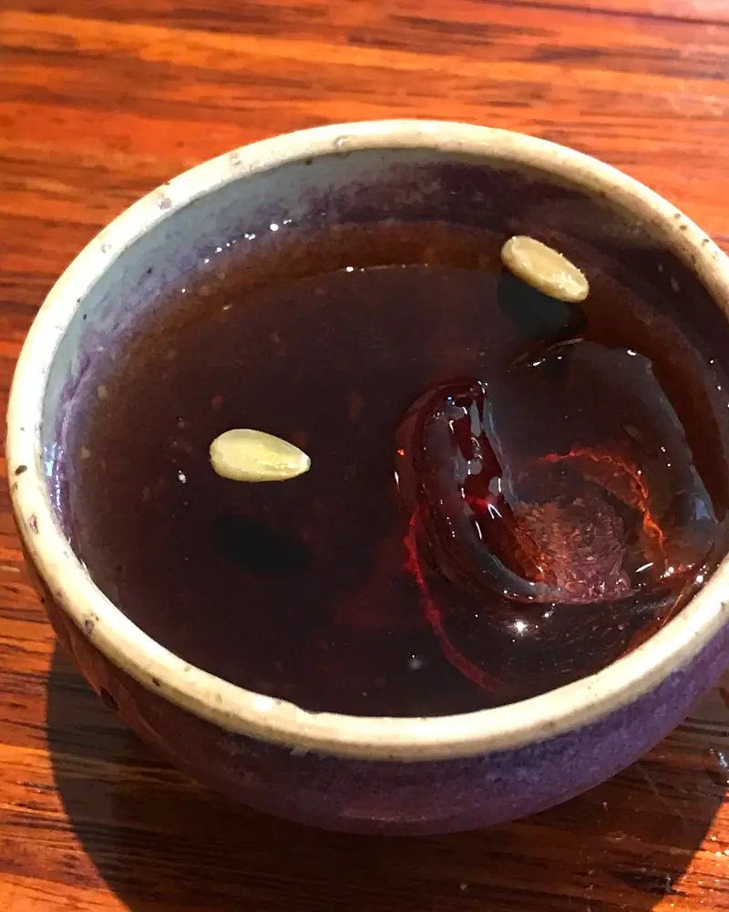 A small ceramic bowl of dark red-brown sujeonggwa with a curl of dried persimmon and a floating pine nut
