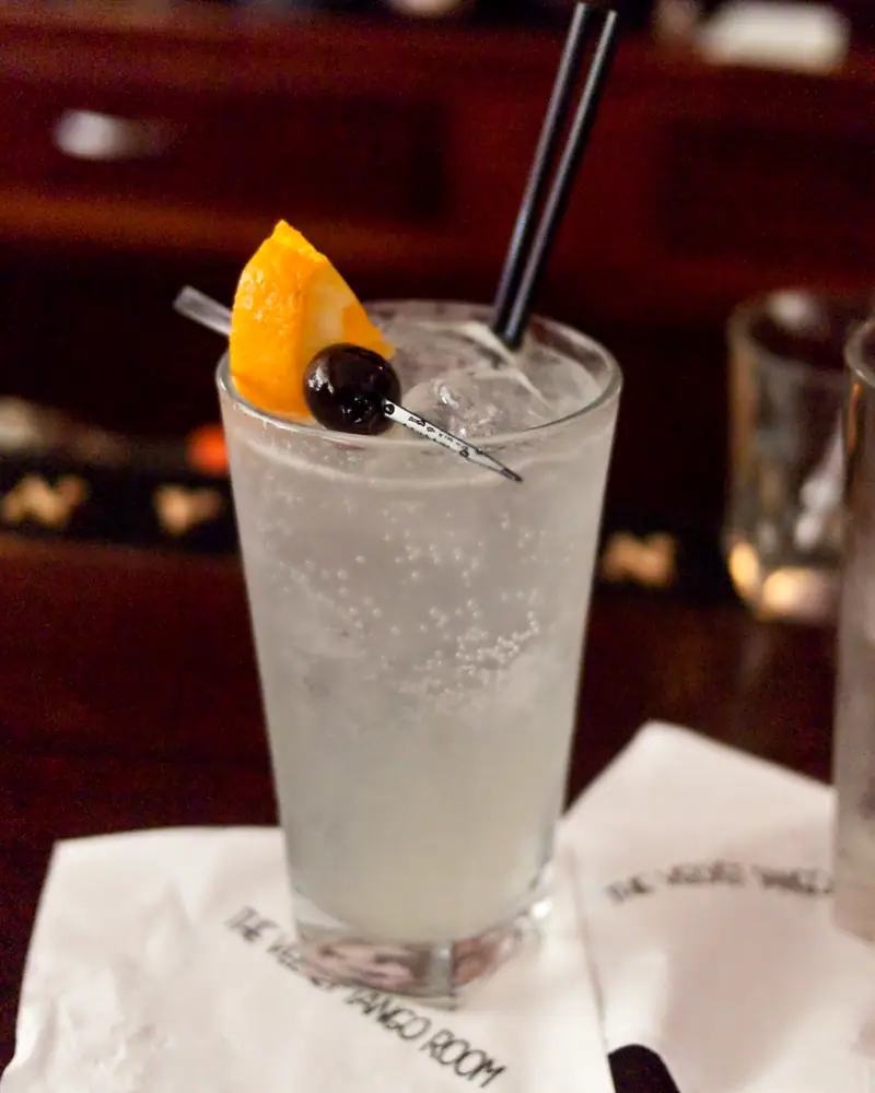 A Tom Collins in a tall glass, pale and fizzing, garnished with an orange wedge and a cherry on a pick, with a straw