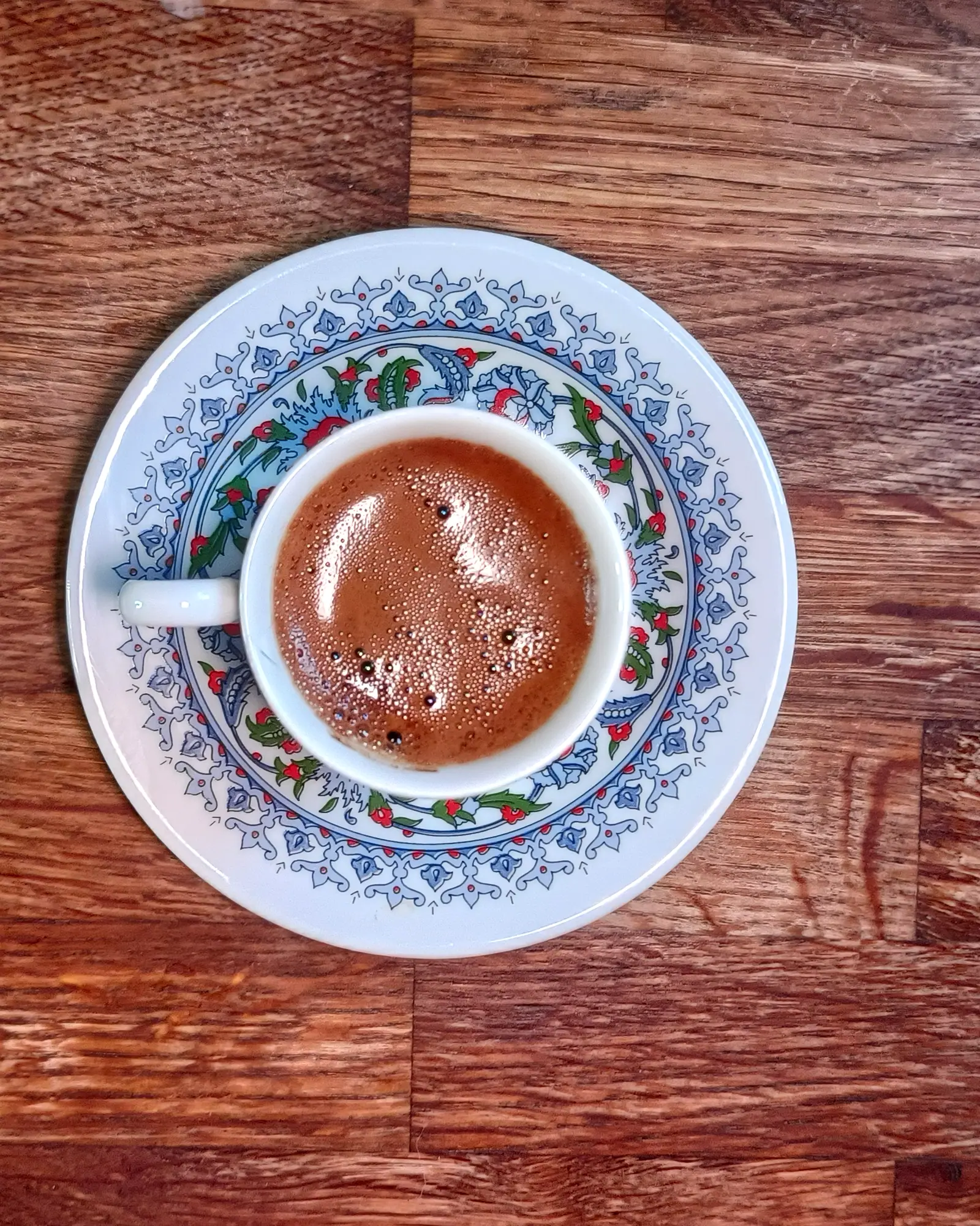 A small cup of Turkish coffee with a thick reddish-brown foam, on a patterned saucer, seen from above