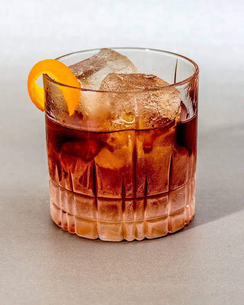 A Vieux Carre served over ice in a rocks glass, deep reddish-amber, with an orange twist against the rim