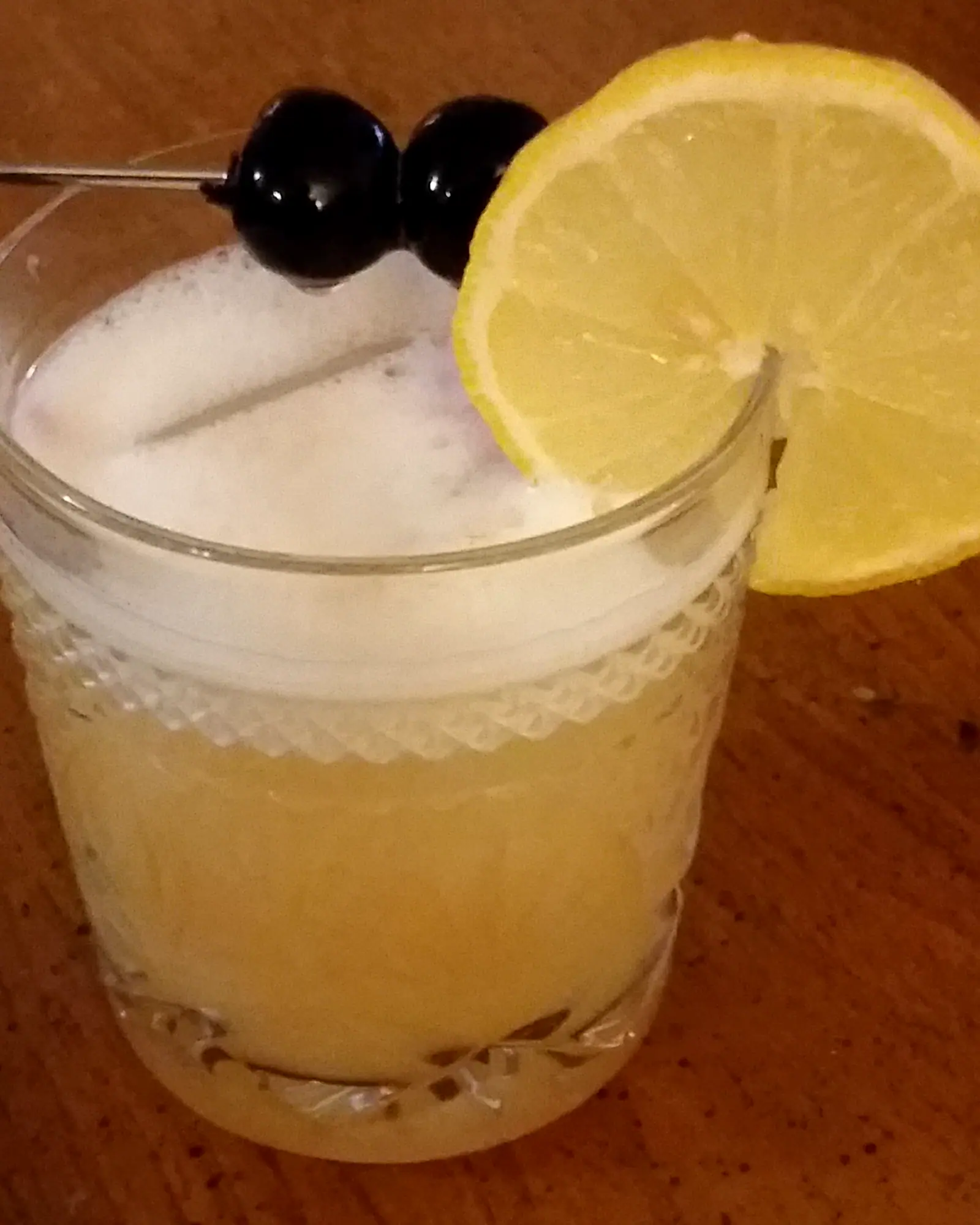 A Whiskey Sour in a heavy tumbler over ice, pale gold under a thin white head, with a lemon wheel and two cherries on a pick